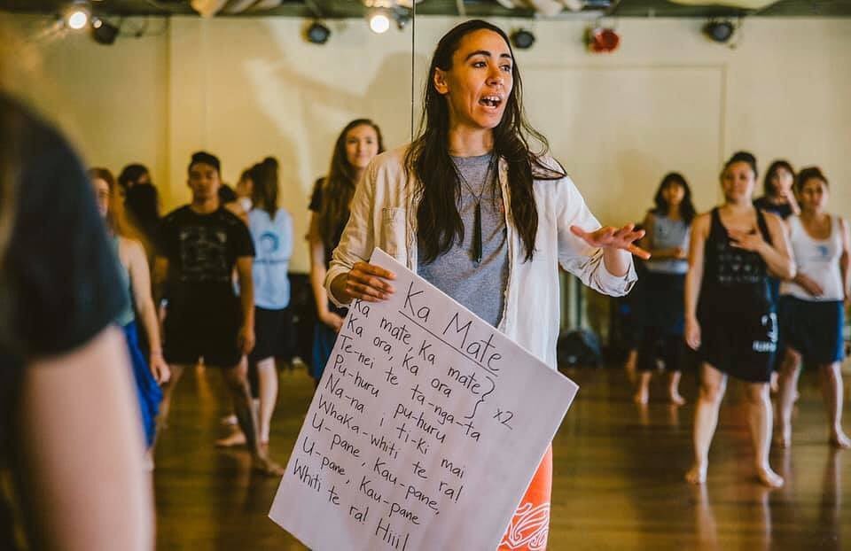 Amelie Butler holding a cue card of traditional songs. She stands in the midst of a full class with all eyes on her. Everyone appears to be listening to Ms. Butler, while holding a pose from a dance.