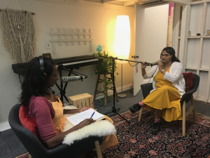 Mrs. Rajamani sits before a microphone in a small studio environment, opposite Ms. Strong who is also at a microphone,